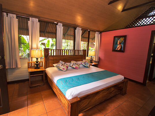 Rustikales Hotelzimmer in Costa Rica mit Holzbetten, farbigen Kissen, rosa Holzwand und Blick ins Grüne.