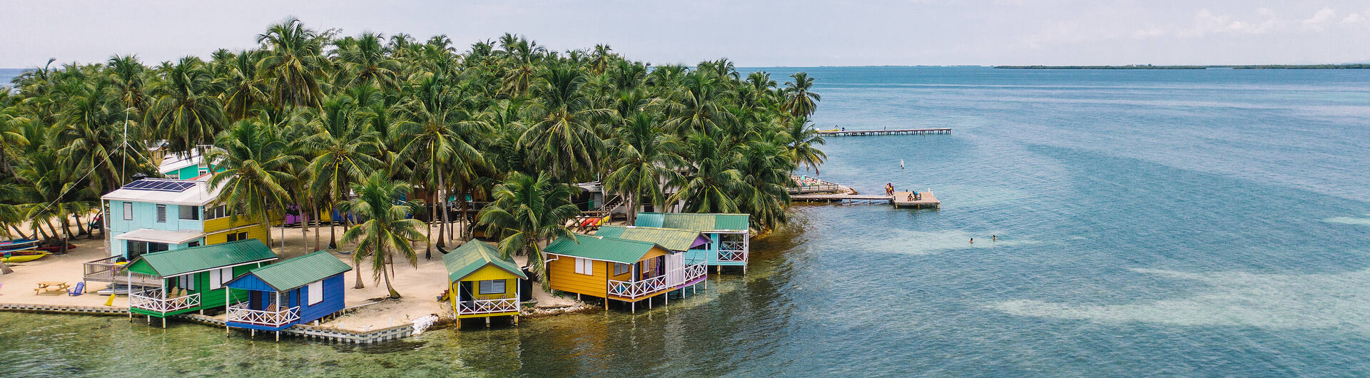 Luftaufnahme der kleinen Insel Tobacco Caye in Belize mit dichtem Palmenbewuchs, bunten Hütten am Sandstrand und umgeben von türkisfarbenem Wasser mit Korallenriffen.