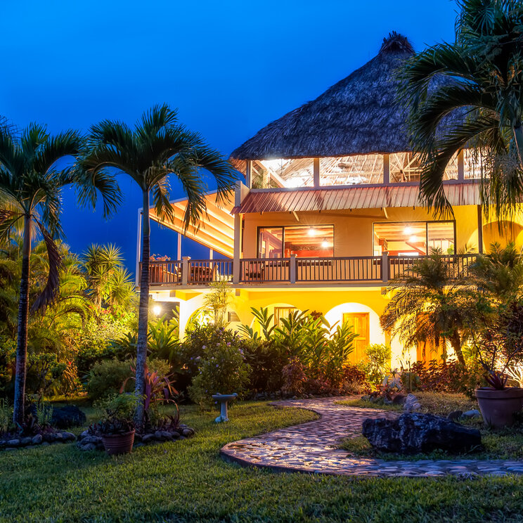 Beleuchtetes Hauptgebäude der Lodge mit Strohdach und Palmen bei Dämmerung in San Ignacio, Belize.