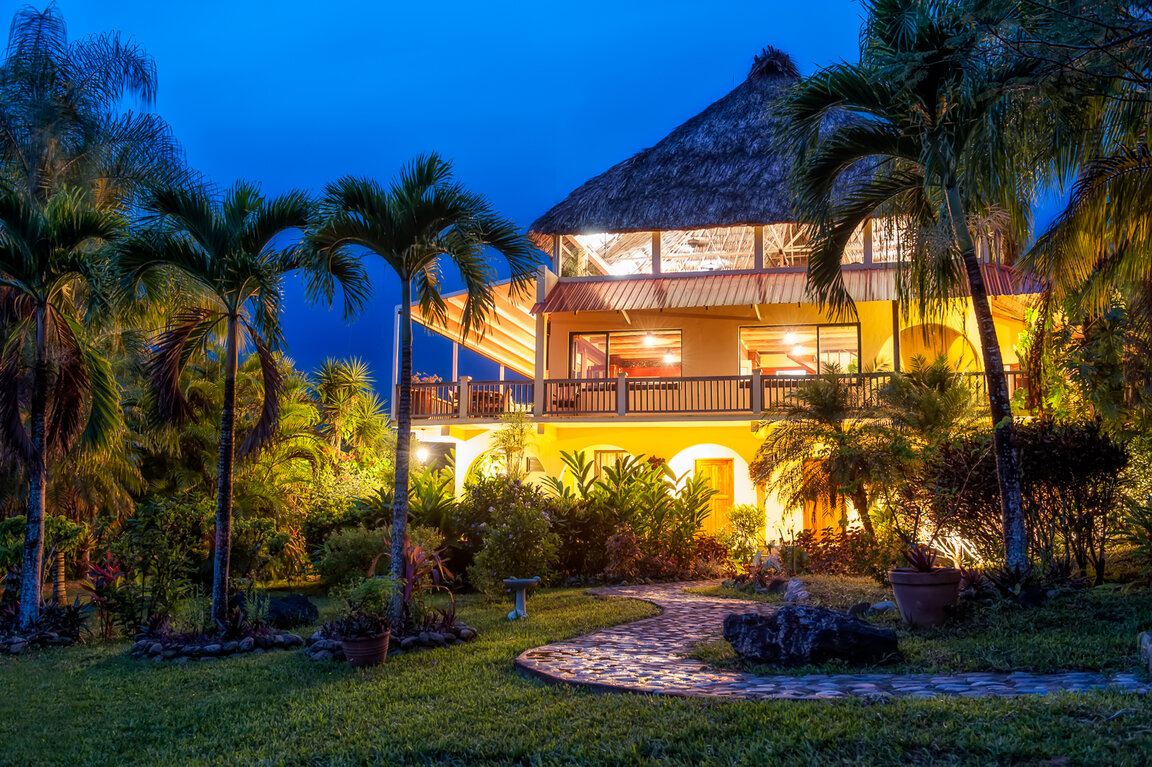 Beleuchtetes Hauptgebäude der Lodge mit Strohdach und Palmen bei Dämmerung in San Ignacio, Belize.