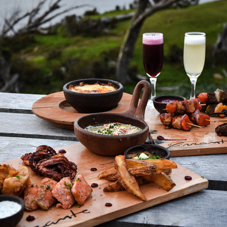 Patagonisches Abendessen auf Holzbrettern mit gegrilltem Fleisch, Beilagen und zwei Gläsern Wein auf einer Terrasse mit Blick ins Grüne.