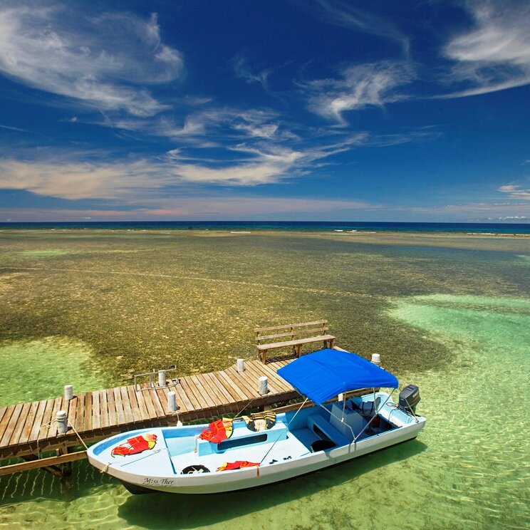 South Water Caye in Belize: Boot am Bootssteg aus Holz mit blauer Himmel und türkisfarbenem Meer im Hintergrund