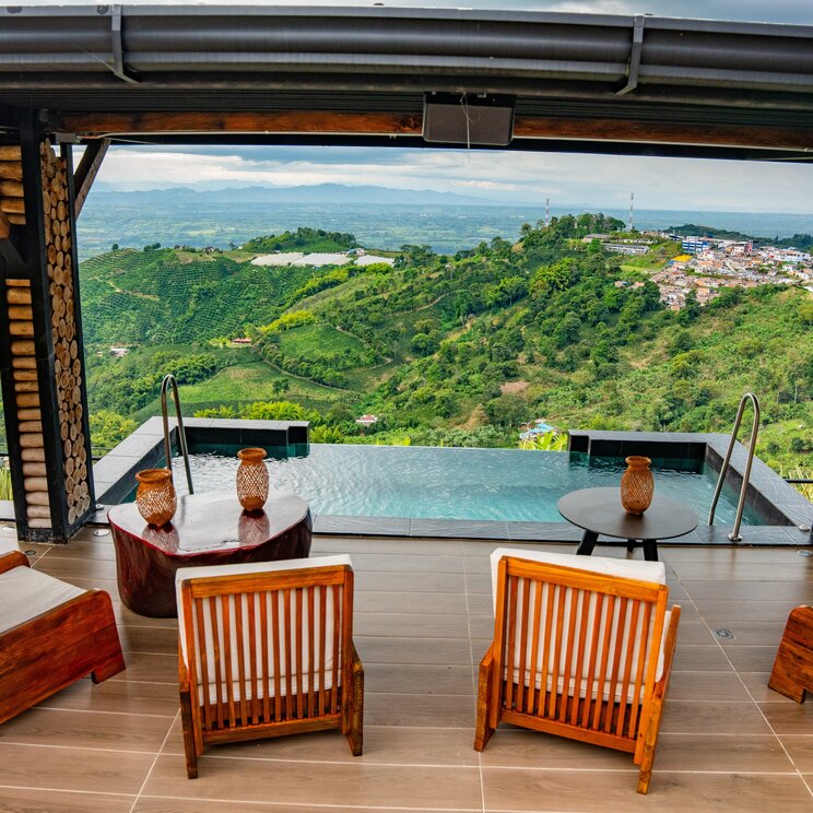 Ueberdachte Lounge mit Holzsesseln, Pool und Weitblick auf gruene Huegel und eine Stadt in Kolumbien.