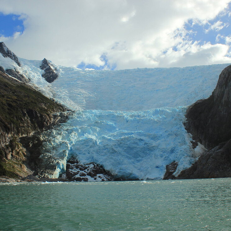 Blick auf den Gletscher Italia auf der Isla Navarino in Patagonien, Chile, mit türkisblauem Eis und umliegenden Felsen, die in ein grünliches Gletscherwasser abfallen.