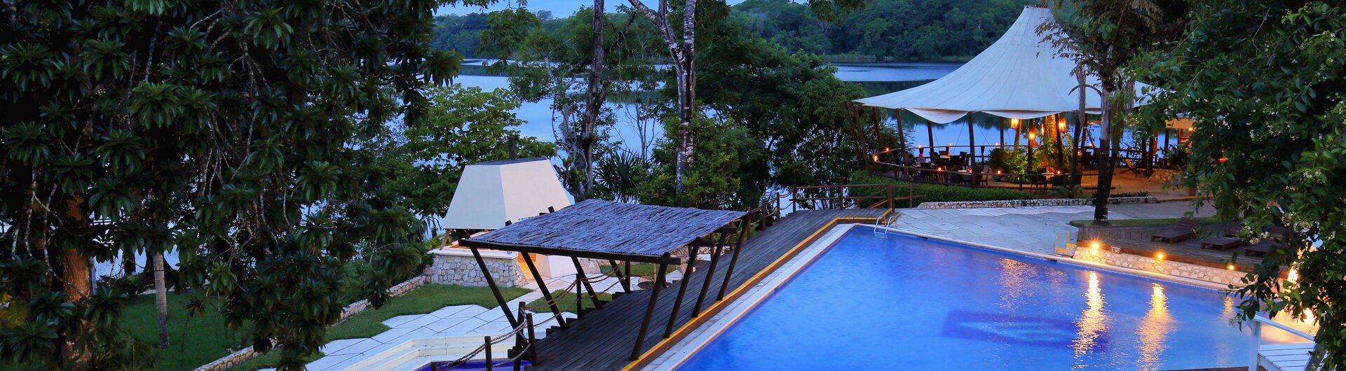 Beleuchteter Aussenpool mit Holzstegen und Sonnensegel in der Daemmerung am See in Guatemala.