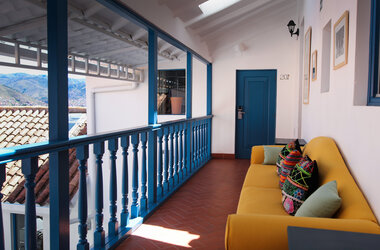 Gelbes Sofa mit bunten Kissen auf einem Balkon mit blauem Holzgelaender und Blick auf Cusco.