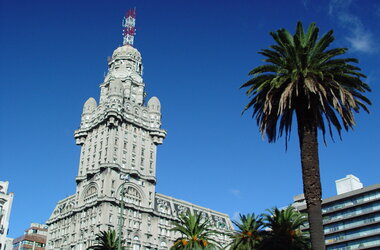 Das markante Hochhaus Palacio Salvo in Montevideo, Uruguay, ragt unter blauem Himmel neben einer Palme in die Höhe.