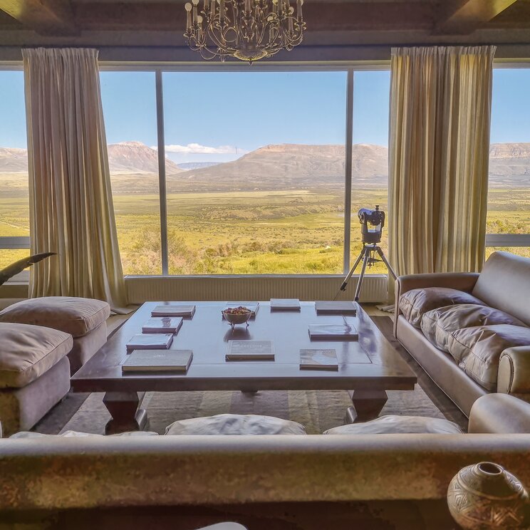 Hotel-Lounge in Patagonien mit Sofas, grossem Couchtisch, Kronleuchter und Panoramafenster mit Blick auf Steppe und Berge.