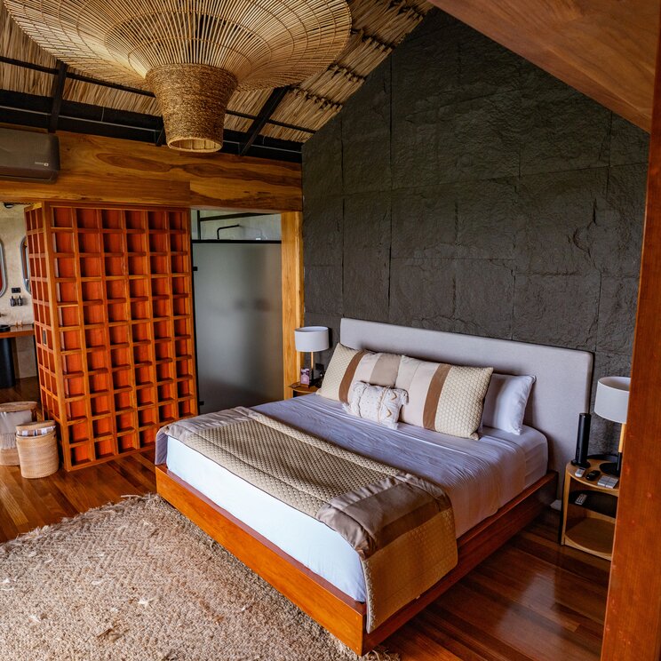 Schlafzimmer mit Doppelbett, Steinwand und Lampenschirm aus geflochtenem Bambus.