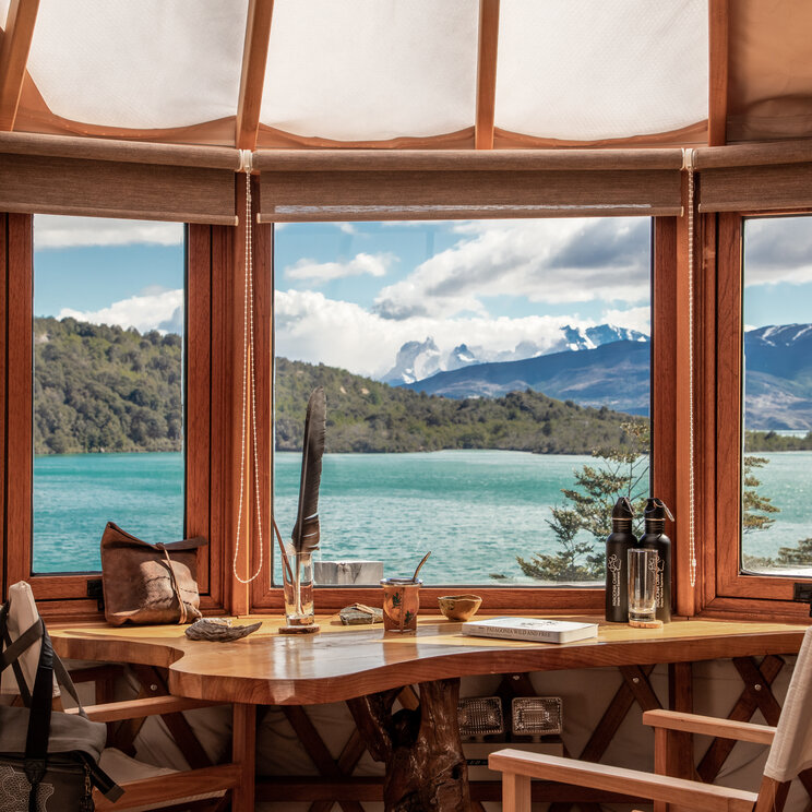 Yurt im Patagonia Camp in Torres del Paine mit Schreibtisch am Panoramafenster, Blick auf türkisfarbenen See und Berglandschaft sowie Holzelementen im Innenraum.