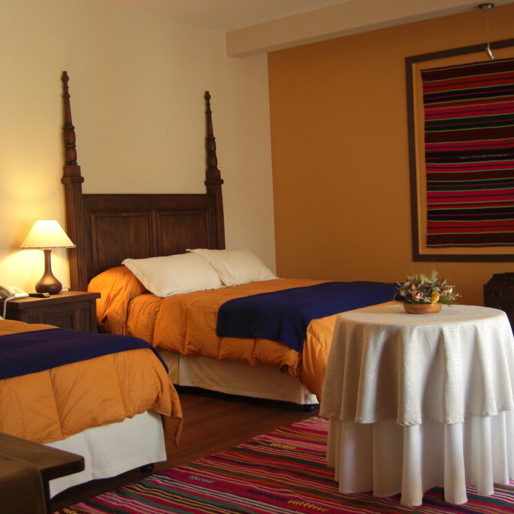 Hotelzimmer in Bolivien mit zwei Betten, orangefarbenen Decken, dunklen Holzpfosten und buntem Wandteppich.