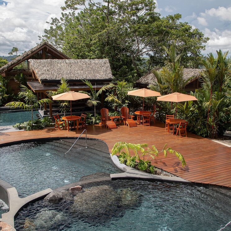 Poolbereich im Hotel Amor Arenal in La Fortuna, Costa Rica, mit organisch geformten Becken, Holzterrasse, Sonnenschirmen und umgeben von tropischer Vegetation.