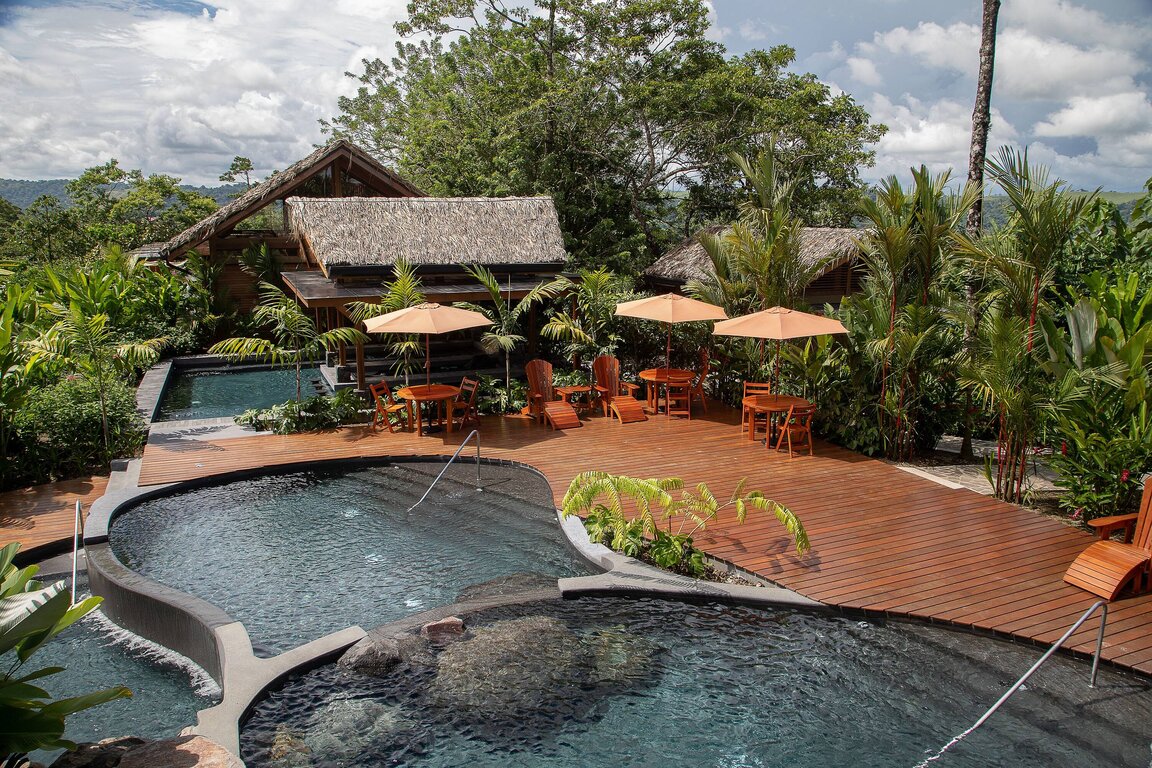 Poolbereich im Hotel Amor Arenal in La Fortuna, Costa Rica, mit organisch geformten Becken, Holzterrasse, Sonnenschirmen und umgeben von tropischer Vegetation.