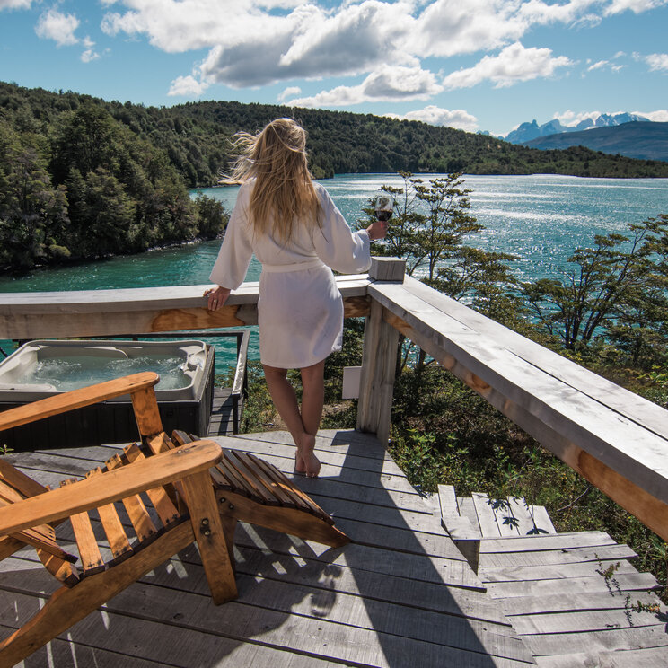 Frau im weißen Bademantel blickt von einer Holzterrasse mit Whirlpool auf den Lago Toro, Chile.