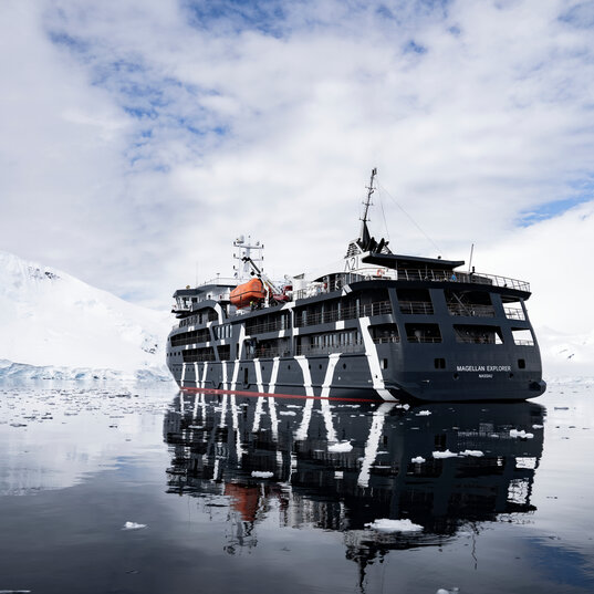 Das Expeditionsschiff Magellan Explorer spiegelt sich im spiegelglatten Wasser mit Treibeis vor Schneebergen in der Antarktis.
