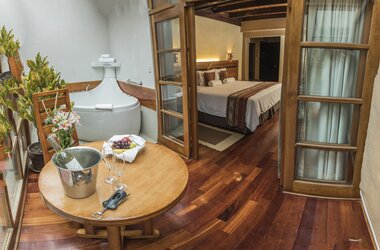 Suite mit Whirlpool, Holztisch mit Weinkühler und Blick durch die geöffnete Tür auf das Doppelbett in Peru.