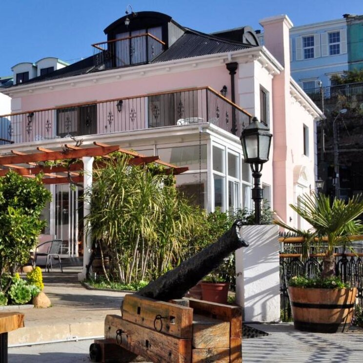 Rosafarbenes Hotelgebaeude mit Terrasse, Pergola und historischer Kanone in Valparaiso, Chile.