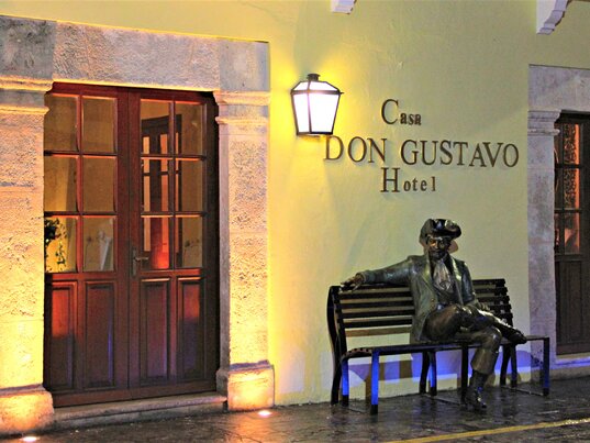 Hoteleingang bei Nacht mit beleuchtetem Schild und Statue auf einer Bank in Campeche, Mexiko.