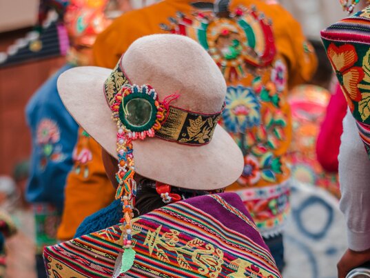 Rueckenansicht einer Person in farbenfroher bolivianischer Tracht mit gemustertem Umhang und hellem Hut.