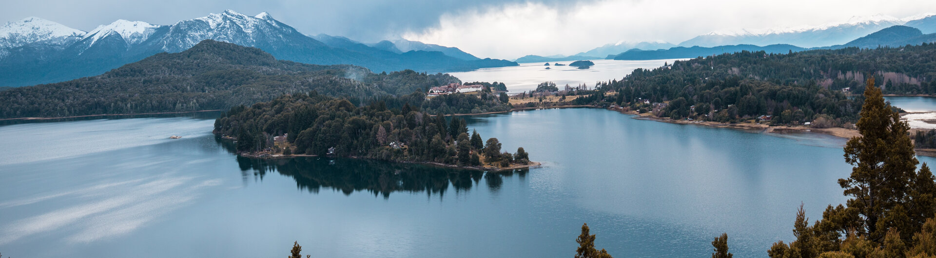 Vogel fliegt ueber dem Nahuel Huapi See und bewaldeten Inseln vor schneebedeckten Bergen in Bariloche, Argentinien.