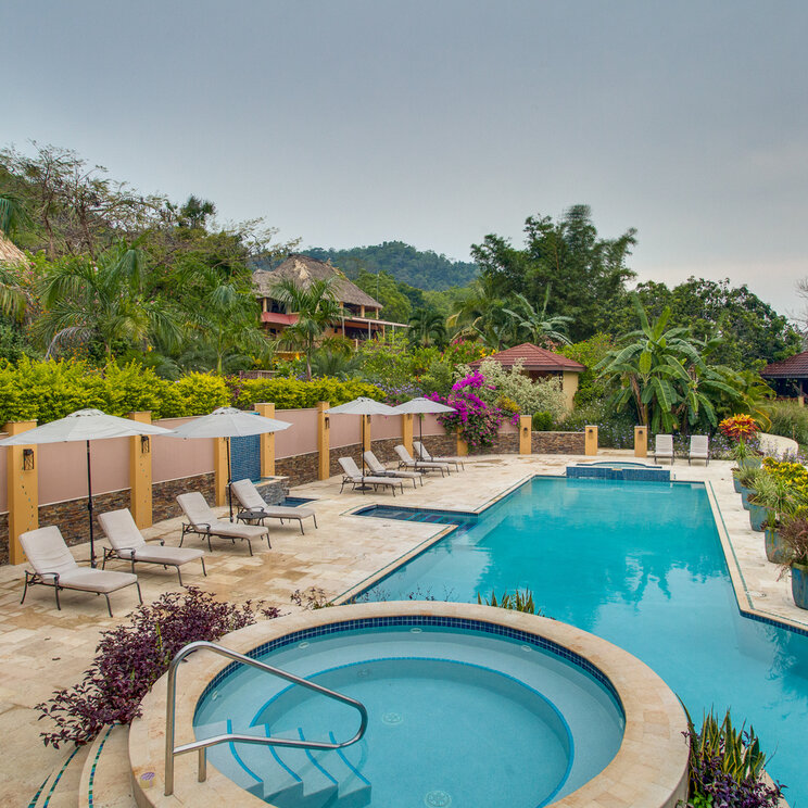 Poolanlage mit Whirlpool, Liegestühlen und Sonnenschirmen vor bewaldeten Hügeln in San Ignacio, Belize.