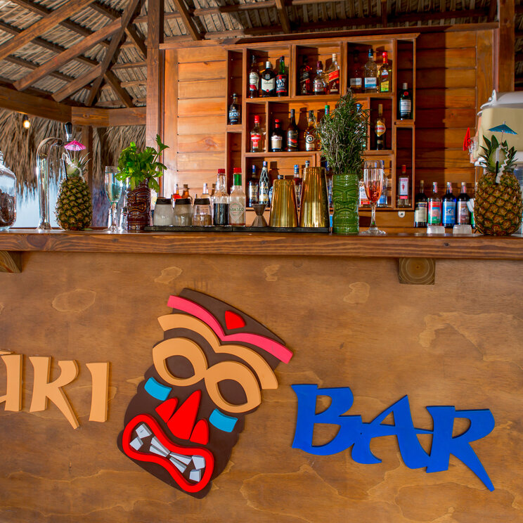 Tiki-Bar aus Holz mit bunten Figuren, Cocktails in Ananas-Bechern und Spirituosen-Regal im Impressive Resort Punta Cana.