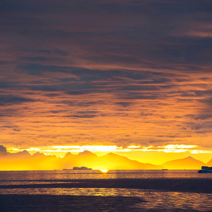 Spektakulärer Sonnenuntergang taucht den Himmel und das Meer über den verschneiten Berggipfeln in glühendes Gold und Orange, Antarktis