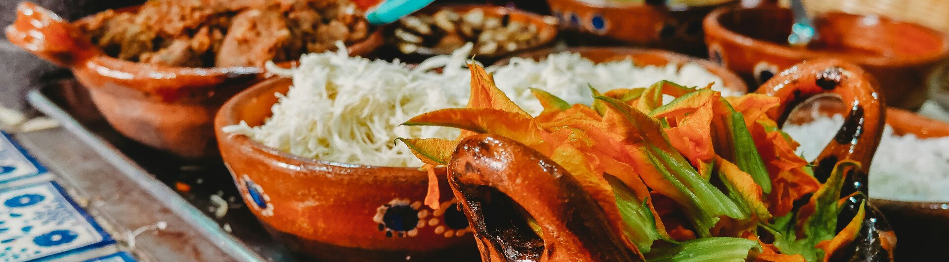 Traditionelle Tonschalen mit Zutaten wie Kürbisblüten, geraspeltem Käse und Fleisch auf blau gefliestem Tisch.