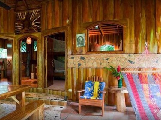 Rustikaler Empfangsbereich aus Holz mit einer bunten Haengematte, Stuhl und geoeffneten Tueren zu Zimmern, Costa Rica.