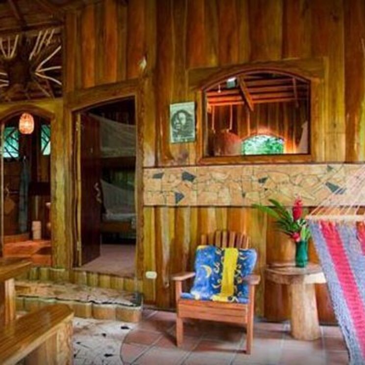 Rustikaler Empfangsbereich aus Holz mit einer bunten Haengematte, Stuhl und geoeffneten Tueren zu Zimmern, Costa Rica.
