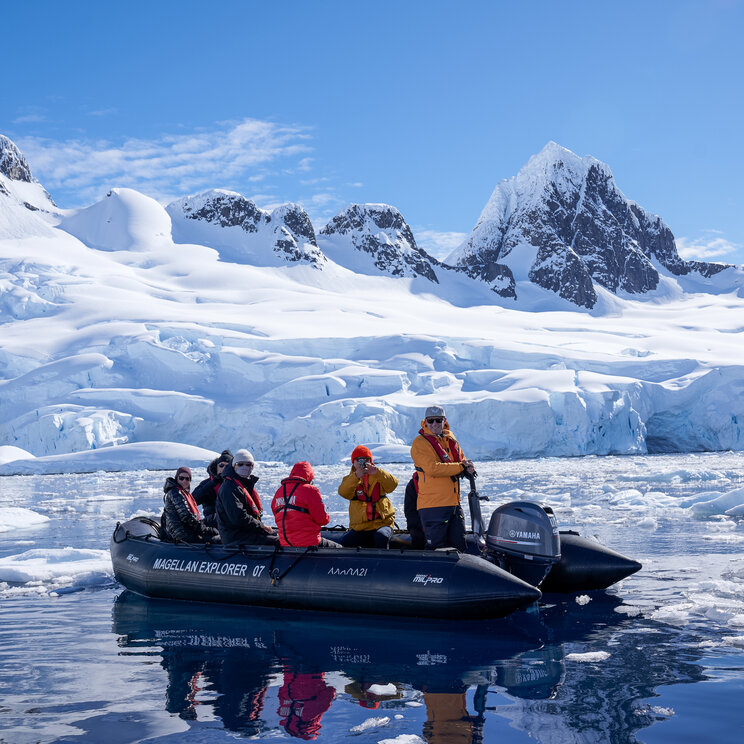 Passagiere in einem Schlauchboot der Magellan Explorer fahren bei blauem Himmel durch Treibeis vor Schneebergen in der Antarktis.