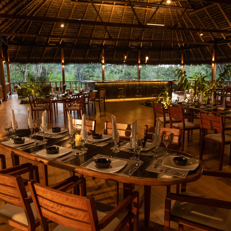 Beleuchtetes Restaurant unter grossem Strohdach mit gedeckten Holztischen in der Sacha Lodge, Ecuador.