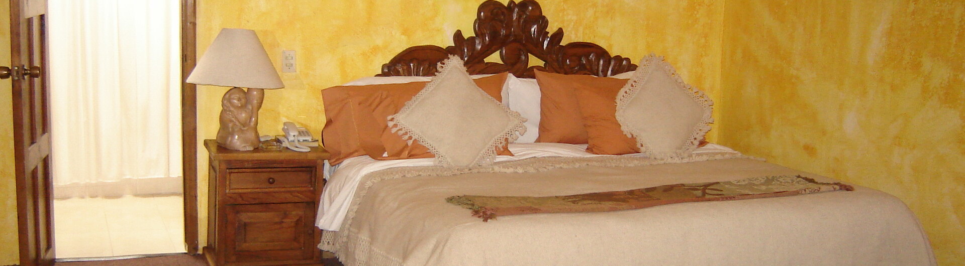 Rustikales Doppelbett mit geschnitztem Kopfteil vor gelber Wand im Hotel Casa Mexicana in San Cristobal, Mexiko.