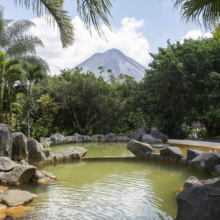 Thermalbecken im Hotel Arenal Paraíso Resort & Spa in La Fortuna, Costa Rica, umgeben von Felsen und tropischer Vegetation mit Blick auf den Vulkan Arenal.