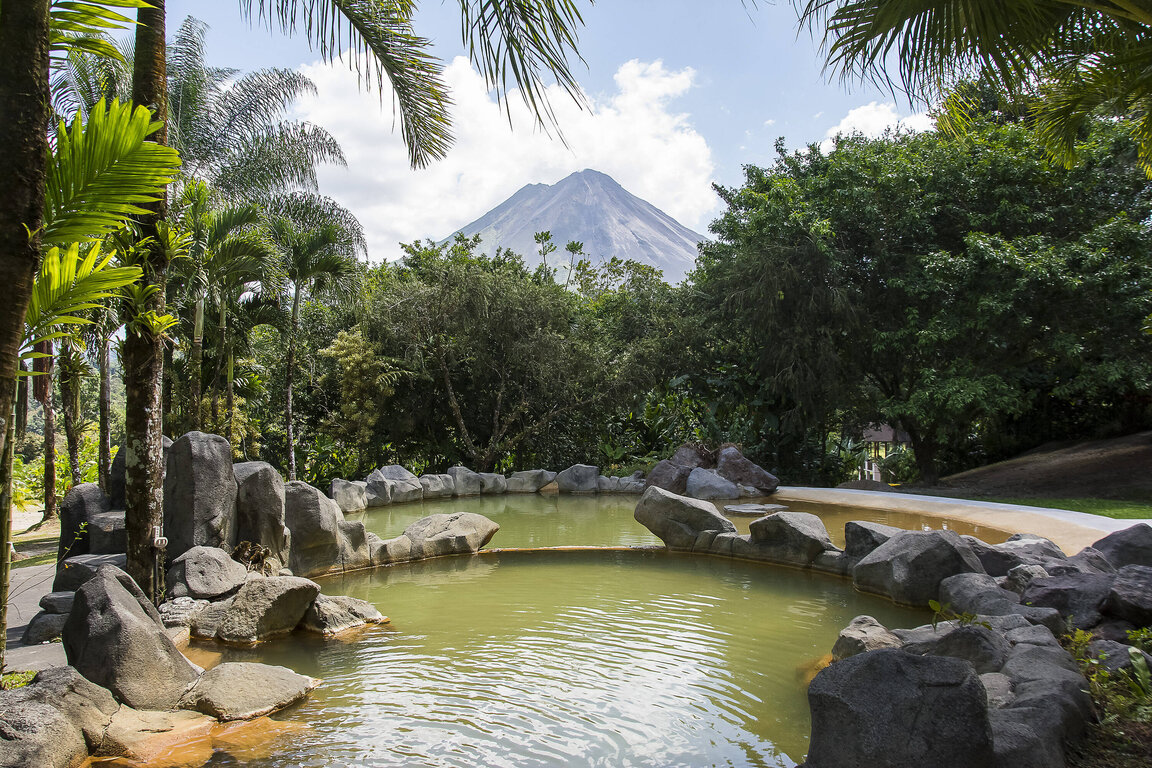 Thermalbecken im Hotel Arenal Paraíso Resort & Spa in La Fortuna, Costa Rica, umgeben von Felsen und tropischer Vegetation mit Blick auf den Vulkan Arenal.