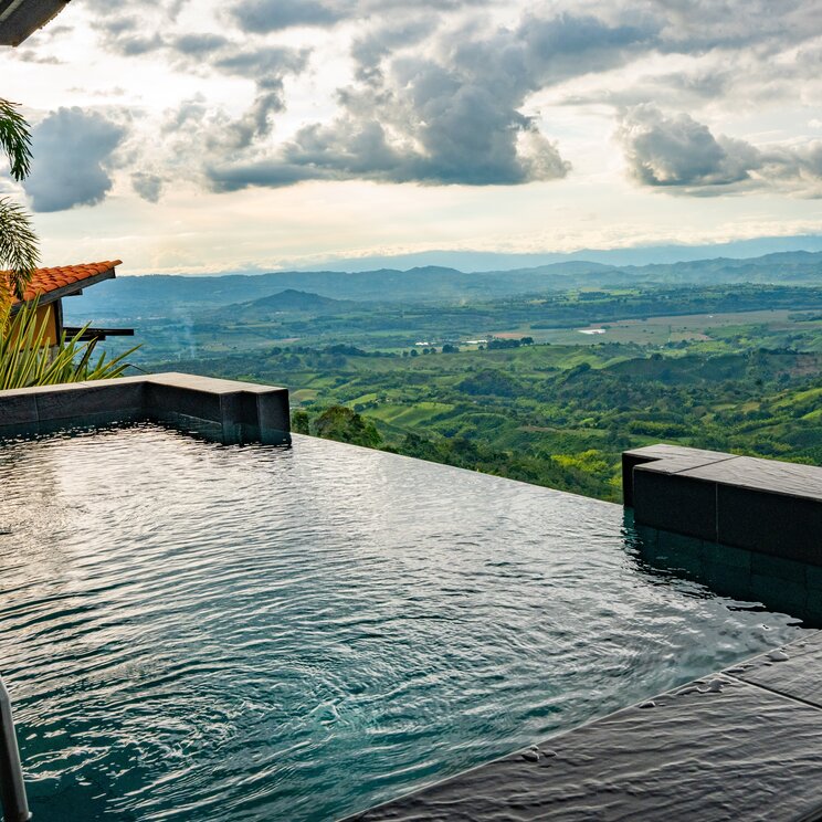 Blick ueber die Kante eines Infinity-Pools auf eine weite, neblige Gebirgslandschaft.