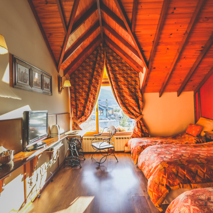 Rustikales Dreibettzimmer im Dachgeschoss des Hotels Tierra de Leyendas in Ushuaia mit hoher Holzverkleidung, drei Einzelbetten mit gemusterten Tagesdecken, Fernseher und einem verzierten Metallstuhl am Fenster, Argentinien.