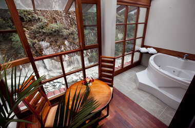 Suite mit Whirlpool, Holztisch und Blick durch große Fenster auf die Steilwand am Fluss in Peru.