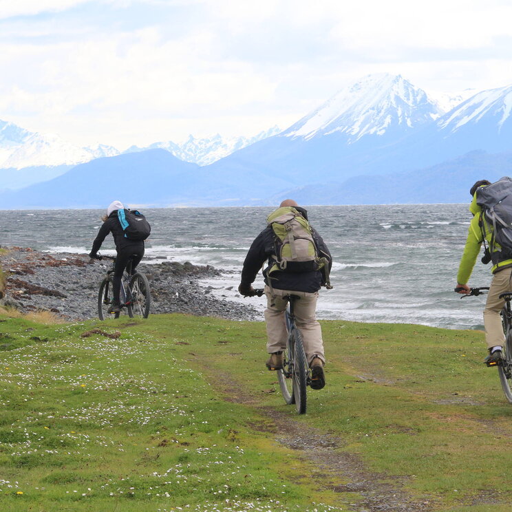 Radfahrer mit Rucksäcken fahren auf Isla Navarino in Patagonien, Chile, entlang der Küste mit Blick auf schneebedeckte Berge.