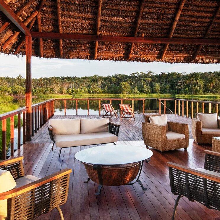 Offener Loungebereich unter Strohdach mit Korbsesseln auf einer Holzterrasse am See, Sacha Lodge