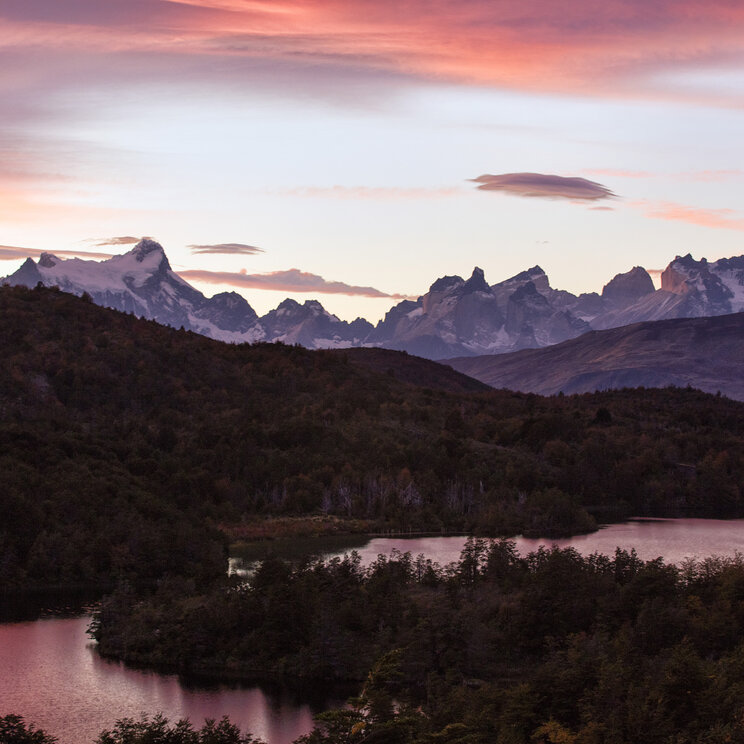 Berglandschaft im Torres del Paine Nationalpark bei Sonnenuntergang mit Seen, bewaldeten Hügeln und markanten Gipfeln der Anden.