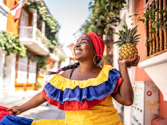 Palenquera in traditioneller gelb-blau-roter Kleidung hält eine Ananas in der Altstadt von Cartagena.