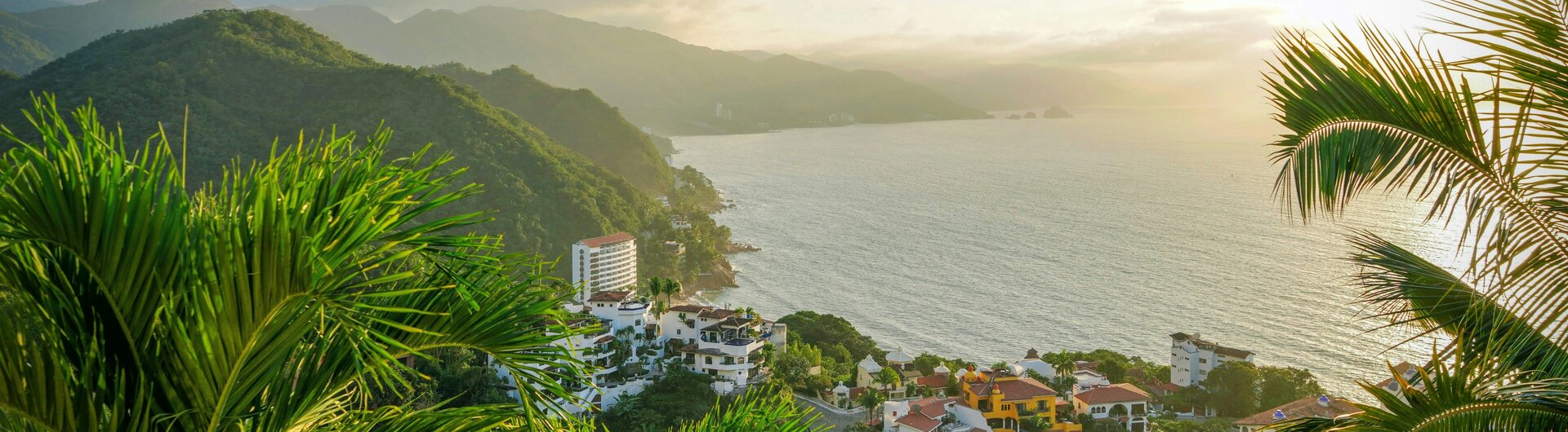 Blick durch Palmwedel auf Hotels an einer bewaldeten Küstenlinie in Puerto Vallarta, Mexiko bei Sonnenuntergang.