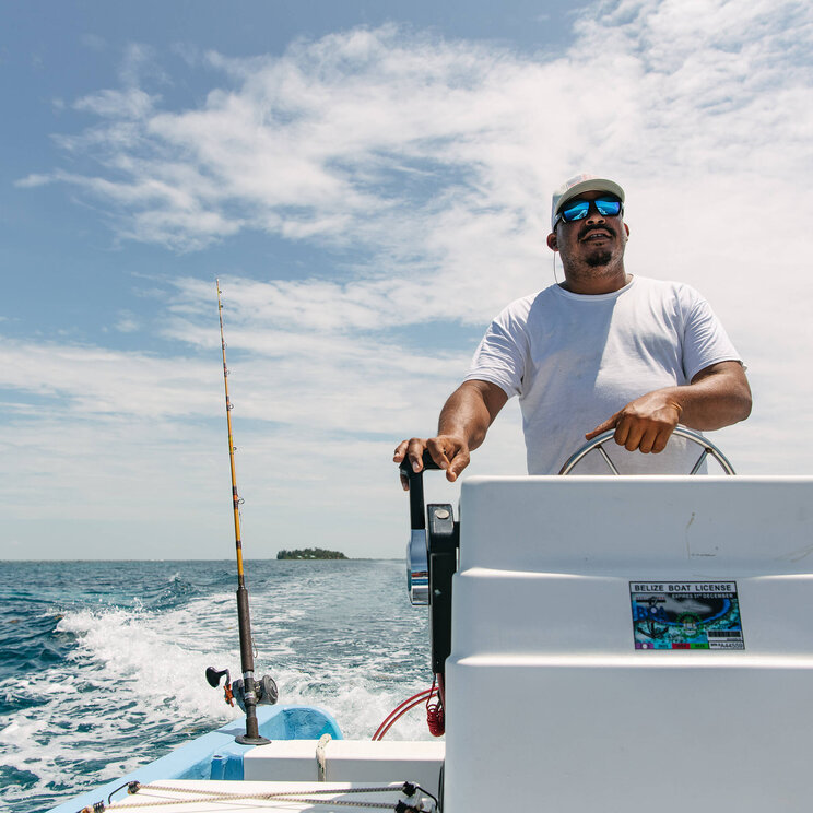 Mann mit Sonnenbrille und Kappe steuert ein Motorboot auf offener See nahe der Tobacco Caye Paradise Lodge in Belize, mit Angelrute im Hintergrund