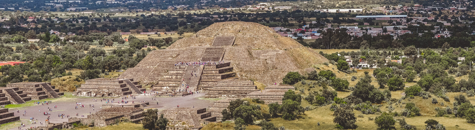 Die Mondpyramide von Teotihuacan in Mexiko mit Besuchern, umgeben von Gruenland und Bergen im Hintergrund.