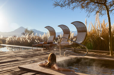 Frau im dampfenden Whirlpool mit Blick auf Liegestühle, Pampasgras und Vulkane in der Atacama-Wüste in Chile.