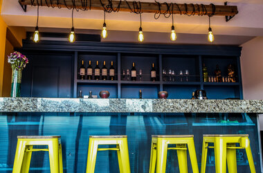 Bar mit Granitplatte, gelben Barhockern und Gluehbirnen an einem Holzbalken vor einem blauen Weinregal.