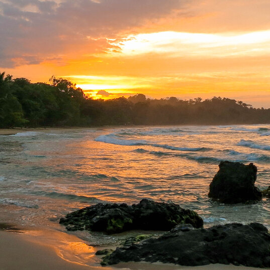 Strand in Bocas del Toro, Panama, mit Wellen, dunklen Felsen im Vordergrund und einem orangefarbenen Sonnenuntergang.