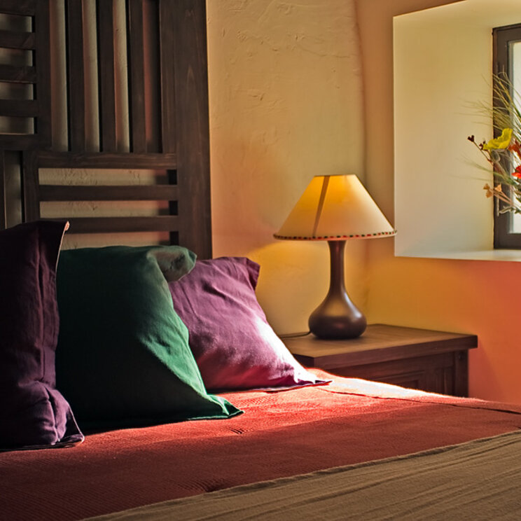 Bett mit dunklem Holz-Kopfteil, bunten Kissen und Blumenstrauß auf dem Fensterbrett in einem Hotel in Sucre.